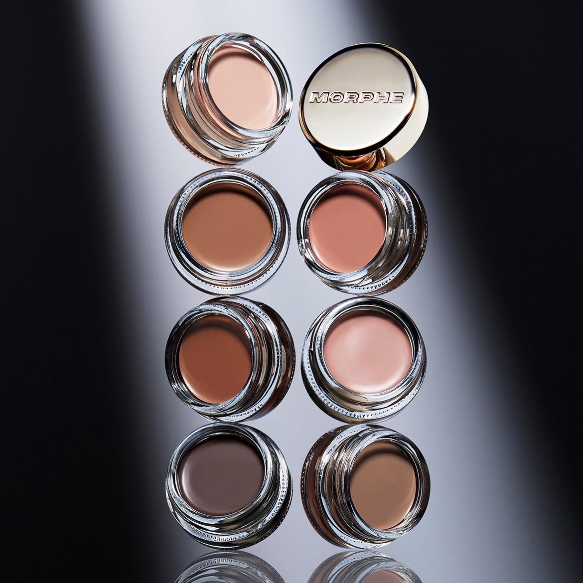 Aquí tienes ocho botes de cristal de Morphe con tapas doradas, cada uno lleno de una crema de diferente tono, desde el beige claro hasta el marrón oscuro, perfectos para definir y dar forma a tus cejas.