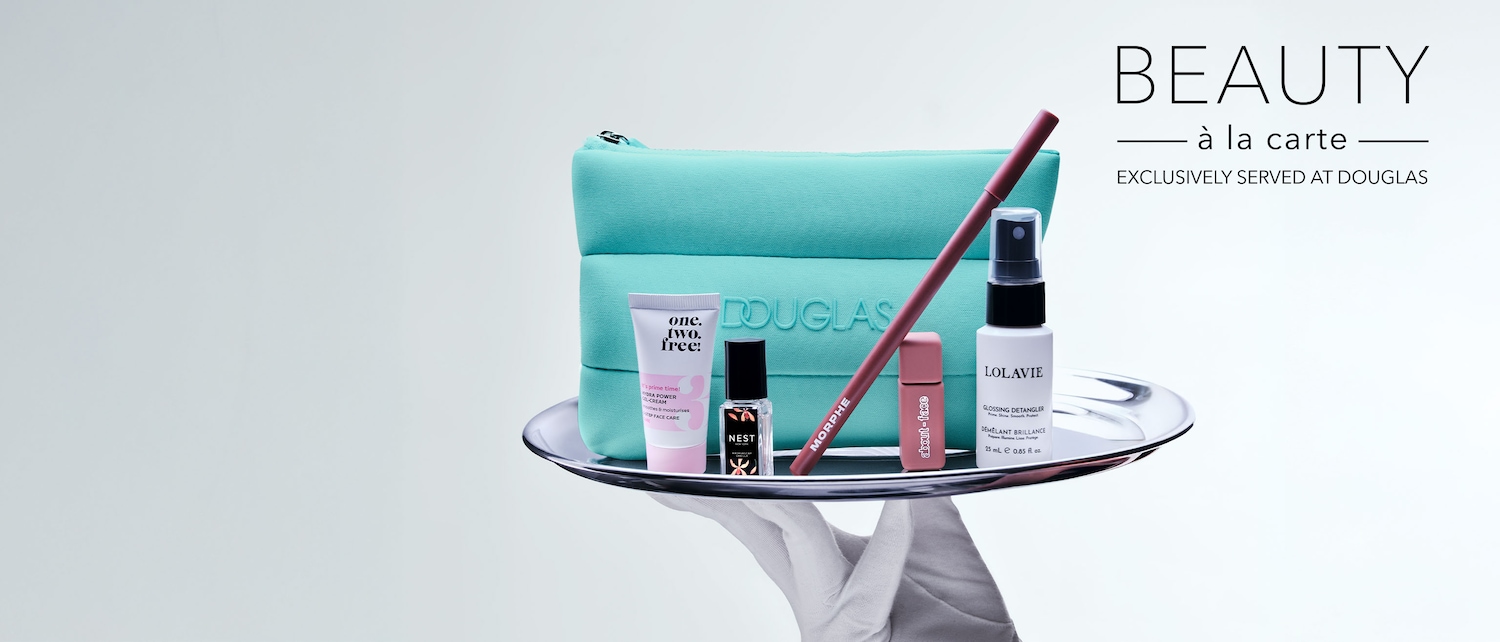 En una bandeja plateada, una mano enguantada presenta un neceser turquesa de Douglas con productos de belleza como crema hidratante, perfume Nest, delineador Morphe, brillo de labios About-face y spray Lolavie, bajo el lema 'Beauty à la carte'.
