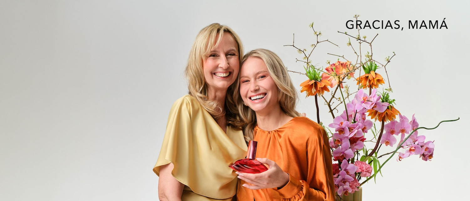 Madre e hija sonriendo, la hija sostiene un frasco de perfume rojo, junto a un arreglo floral y el texto 'Gracias, Mamá'.