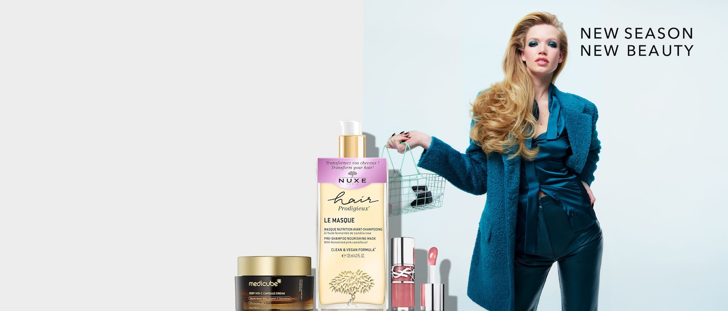 Una mujer rubia con un abrigo azul y pantalones de cuero sostiene una cesta de la compra con productos de belleza, incluyendo una crema Medicube Deep Vita C Capsule, una mascarilla capilar Nuxe Hair Prodigieux y un brillo de labios rosa.