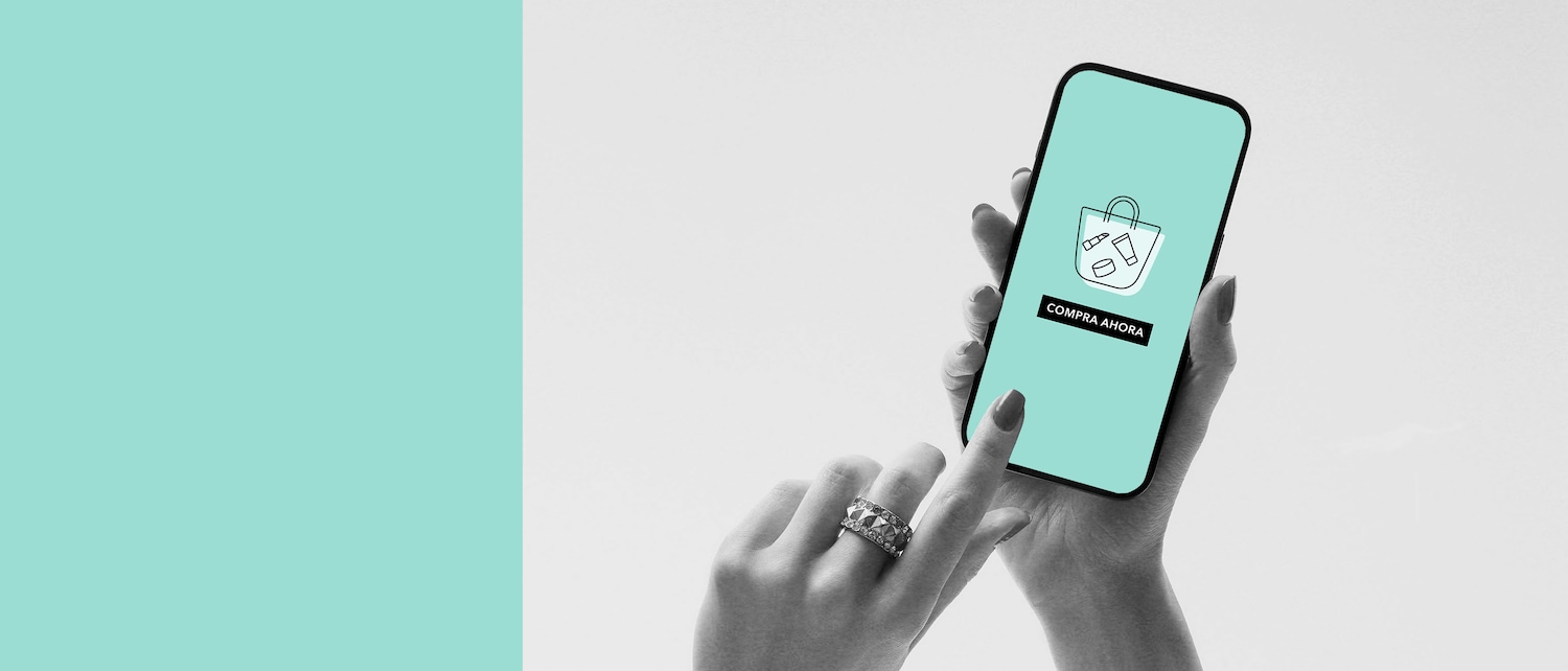 En la imagen en blanco y negro, una mano sostiene un teléfono con una pantalla color aguamarina que muestra un icono de una bolsa de compras y el texto "Compra Ahora".