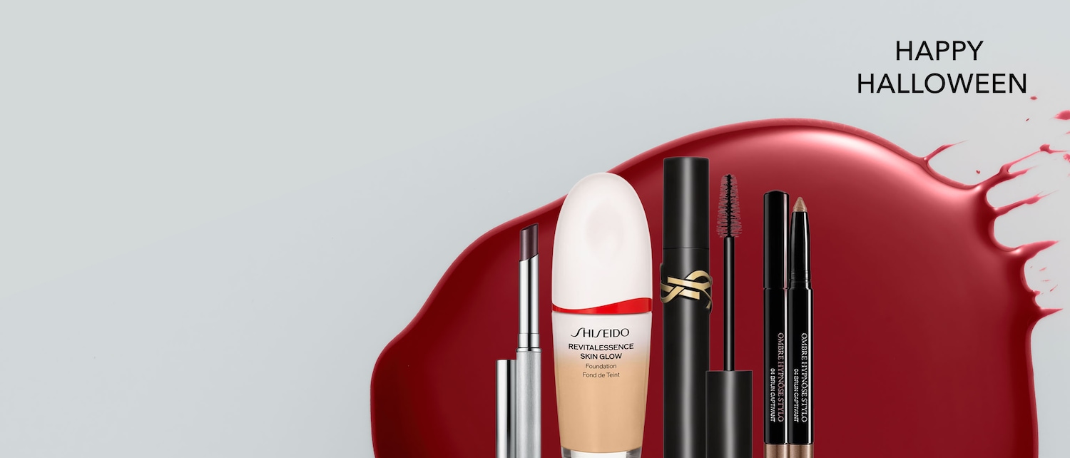 Conjunto de maquillaje que incluye base Shiseido, lápiz labial oscuro, máscara negra y sombra de ojos en barra, presentado sobre un fondo gris con una mancha roja y el texto Feliz Halloween. ¡Compra ahora y completa tu look!