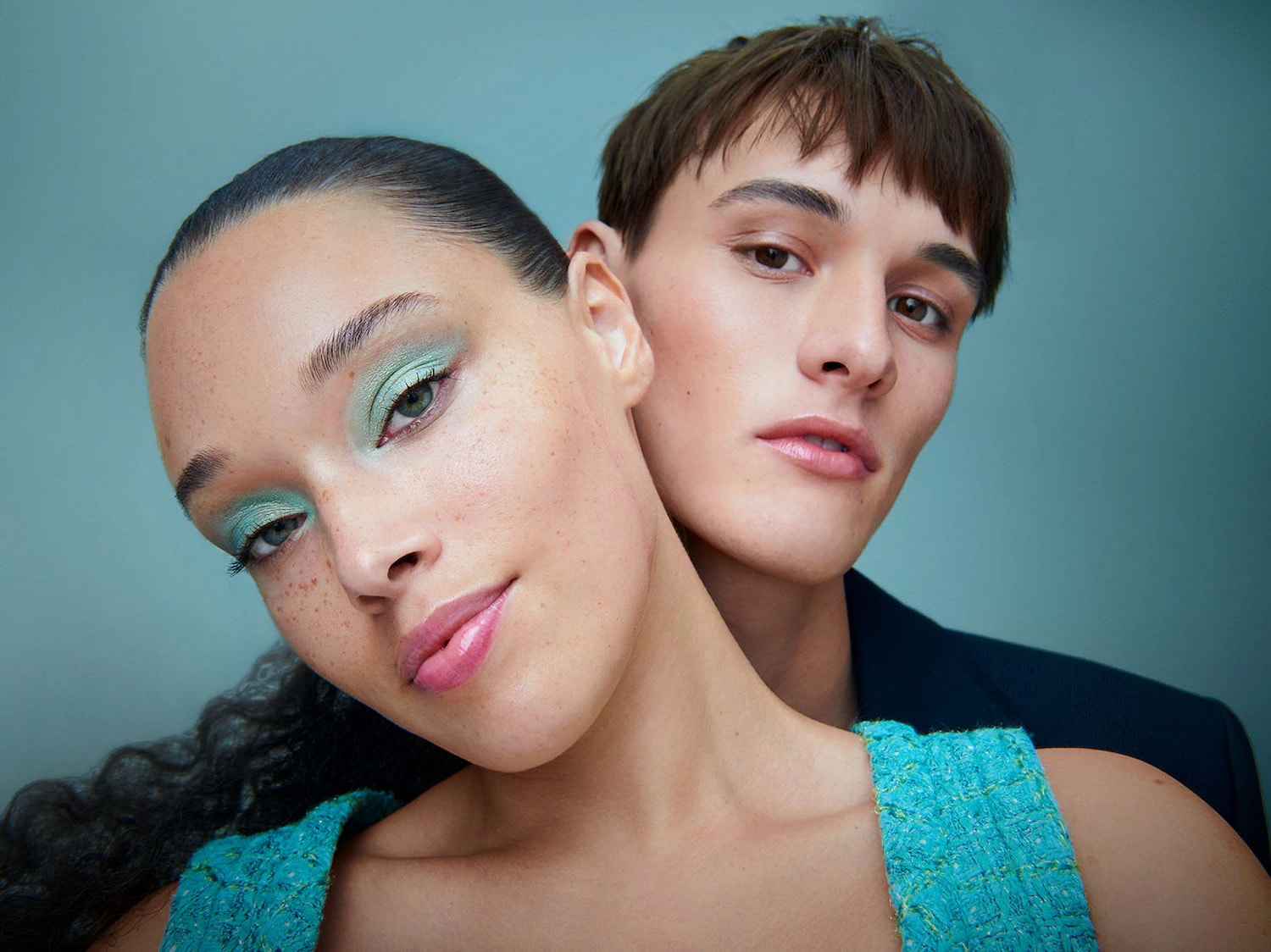 Una mujer con sombra de ojos verde menta y labios rosados brillantes, con un hombre detrás de ella, ambos con un aspecto fresco y natural.